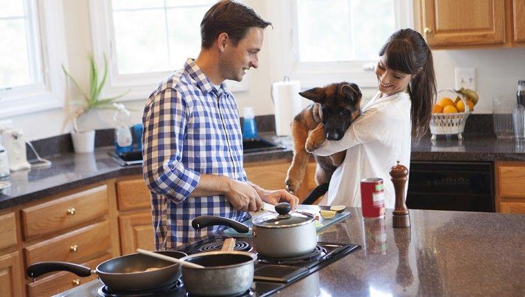 Couple in the kitchen cooking together . Women holding their dog in hands and he looking at food